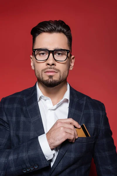 Bearded businessman in plaid blazer and glasses putting credit card in pocket on red — Stock Photo