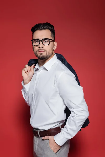 Hombre de negocios barbudo con camisa blanca y gafas que sostienen la chaqueta y posan con la mano en el bolsillo en rojo - foto de stock