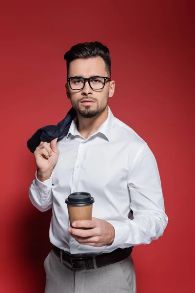 Hombre de negocios barbudo con camisa blanca y gafas que sostienen la chaqueta y la taza de papel en rojo - foto de stock