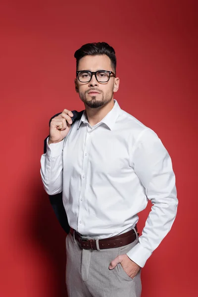 Молодой бизнесмен в белой рубашке и очках, держащий блейзер и позирующий с рукой в кармане на красном — стоковое фото