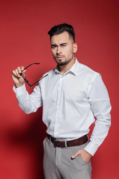 Hombre de negocios barbudo con camisa blanca sosteniendo gafas y posando con la mano en el bolsillo en rojo - foto de stock