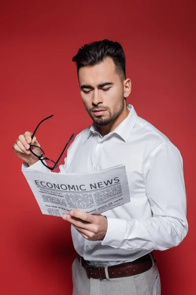 Hombre de negocios barbudo con camisa blanca sosteniendo gafas y leyendo noticias sobre rojo - foto de stock