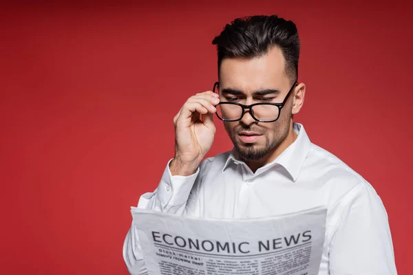 Hombre de negocios barbudo con camisa blanca ajustando gafas y leyendo noticias sobre rojo - foto de stock
