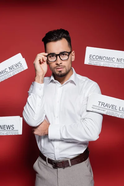 Bearded businessman in white shirt adjusting glasses near newspapers on red — Stock Photo
