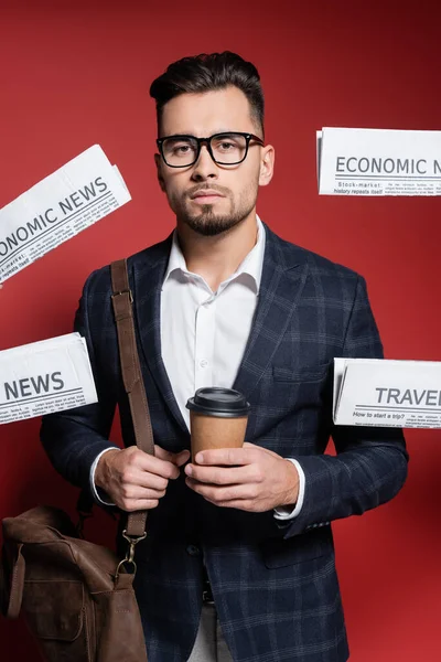 Bearded businessman in glasses and suit holding paper cup near newspapers on red — Stock Photo