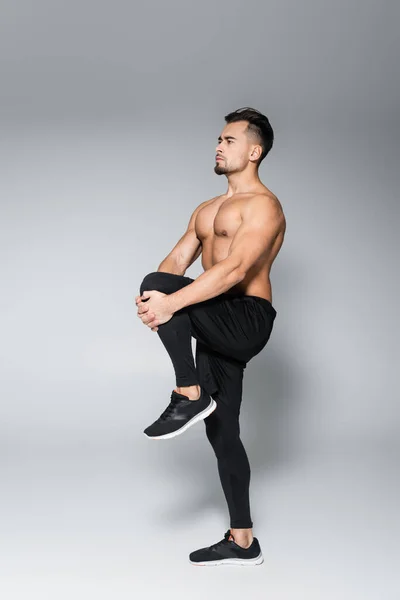 Full length of young muscular sportsman stretching leg on grey — Stock Photo