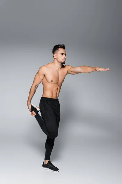 Full length of young shirtless sportsman stretching leg and standing with outstretched hand on grey — Stock Photo