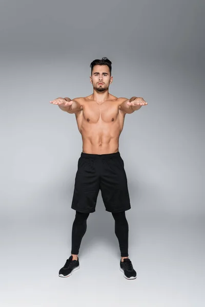 Full length of young shirtless sportsman exercising with outstretched hands on grey — Stock Photo