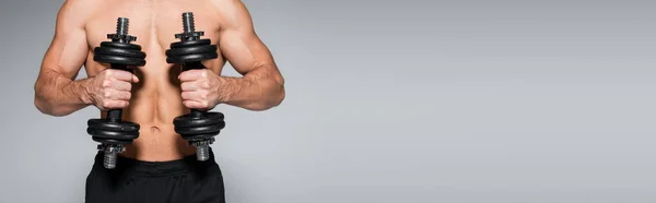 Частичный вид безрубашечного и мускулистого спортсмена, работающего с гантелями, изолированными на сером, баннере — стоковое фото