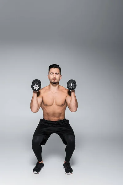 Full length of strong sportsman doing squat with dumbbells on grey — Stock Photo