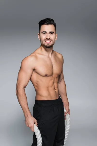 Cheerful and shirtless sportsman holding battle ropes on grey — Stock Photo