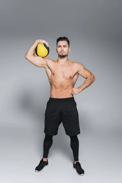 Longitud completa del deportista sin camisa y barbudo sosteniendo la pelota y de pie con la mano en la cadera en gris - foto de stock