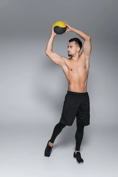 Longitud completa del deportista sin camisa y barbudo sosteniendo la pelota por encima de la cabeza en gris - foto de stock