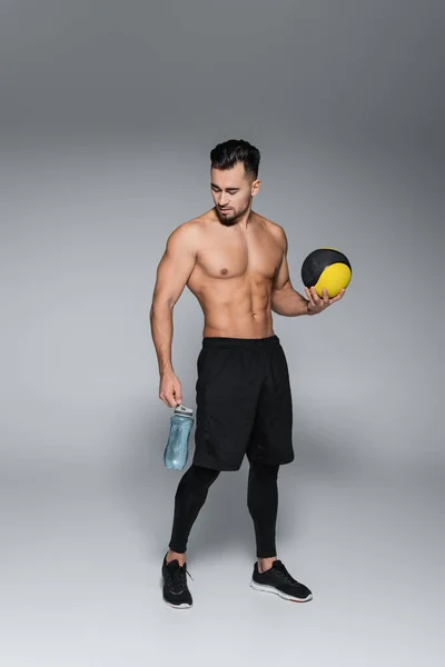 Tutta la lunghezza dello sportivo senza maglietta e barbuto che tiene palla e bottiglia sportiva sul grigio — Foto stock