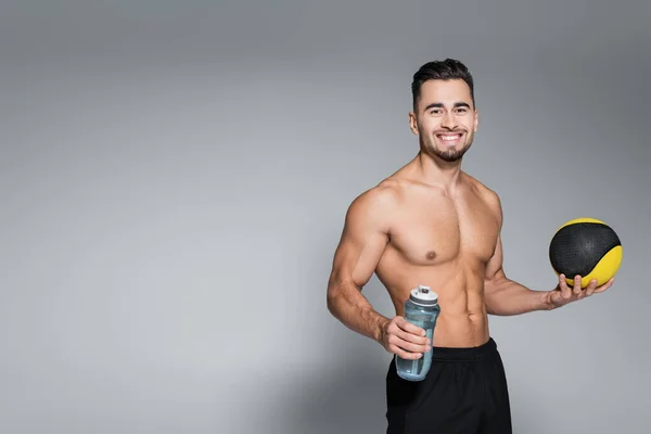 Без сорочки і усміхнений спортсмен тримає м'яч і спортивну пляшку на сірому — стокове фото