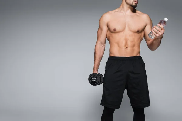 Cropped view of shirtless sportsman holding dumbbell and bottle with water on grey — Stock Photo