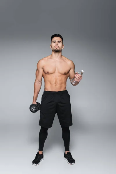 Full length of shirtless sportsman holding dumbbell and bottle with water on grey — Stock Photo