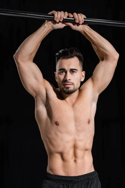 Hemdloser und muskulöser Sportler, der in die Kamera in der Nähe des horizontalen Balkens schaut, isoliert auf schwarz — Stockfoto