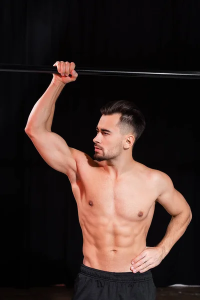 Sem camisa e desportista muscular olhando para longe e posando com a mão no quadril perto da barra horizontal isolada no preto — Fotografia de Stock