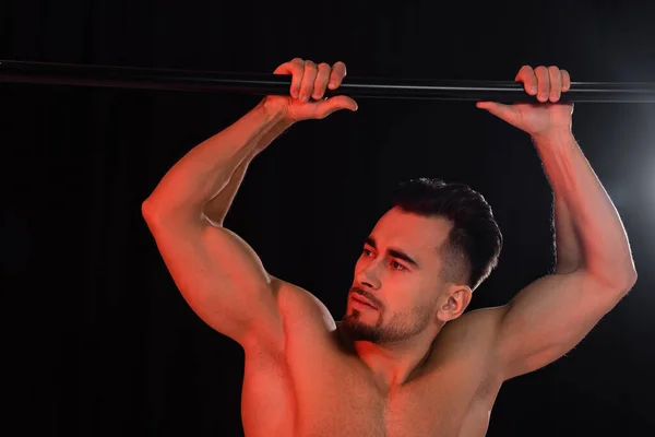 Red lighting on muscular sportsman looking away and leaning on horizontal bar isolated on black — Stock Photo