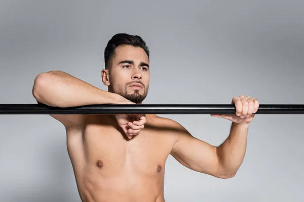 Muscular sportsman looking away and leaning on horizontal bar isolated on grey — Stock Photo