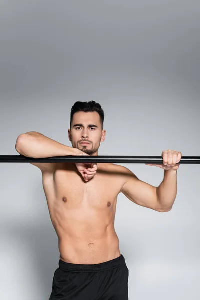 Muscular sportsman looking at camera and leaning on horizontal bar on grey — Stock Photo