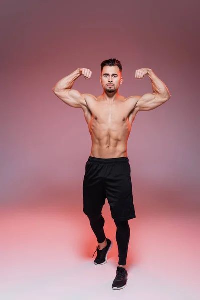 Full length of strong man showing muscles and posing on pink and grey — Stock Photo