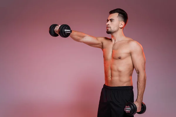 Shirtless man training with dumbbells and looking away on pink and grey — Stock Photo