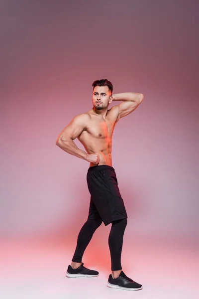 Full length of shirtless sportsman posing while showing muscles on grey and pink — Stock Photo