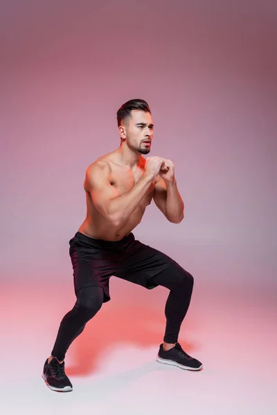 Comprimento total de desportista sem camisa fazendo exercício agachamento em cinza e rosa — Fotografia de Stock