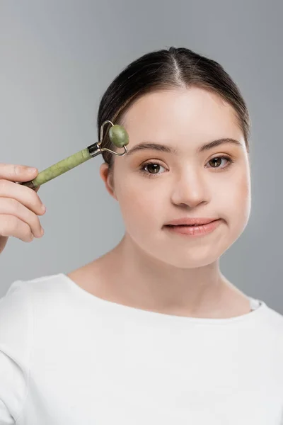 Young woman with down syndrome holding jade roller and looking at camera isolated on grey — Stock Photo