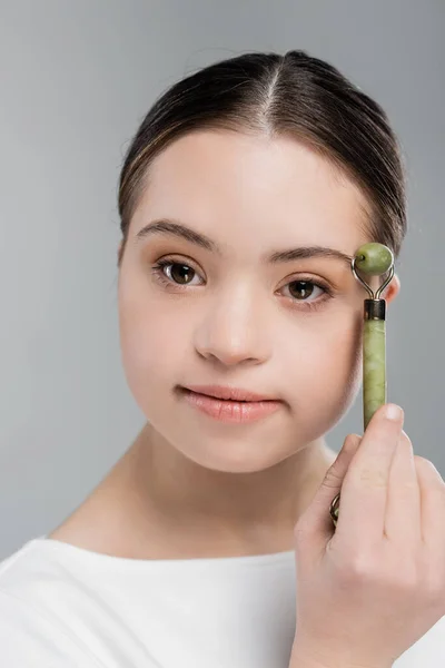 Young woman with down syndrome massaging face with jade roller isolated on grey — Stock Photo
