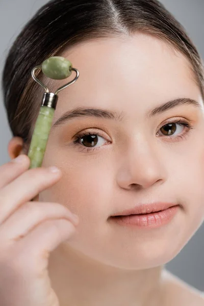 Young woman with down syndrome holding blurred jade roller isolated on grey — Stock Photo