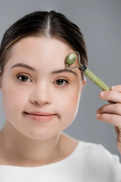 Junge Frau mit Down-Syndrom massiert Stirn mit Jade-Rolle isoliert auf grau — Stockfoto
