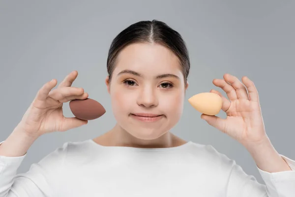 Young woman with down syndrome holding beauty blenders isolated on grey — Stock Photo