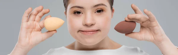 Young woman with down syndrome holding beauty blenders and looking at camera isolated on grey, banner — Stock Photo