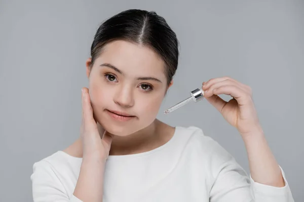 Woman with down syndrome touching face and holding pipette of serum isolated on grey — Stock Photo