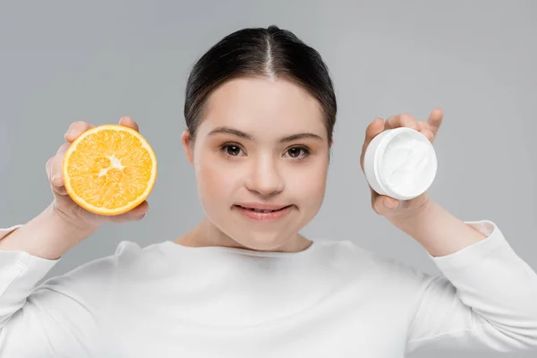 Smiling woman with down syndrome holding cream and orange isolated on grey — Stock Photo