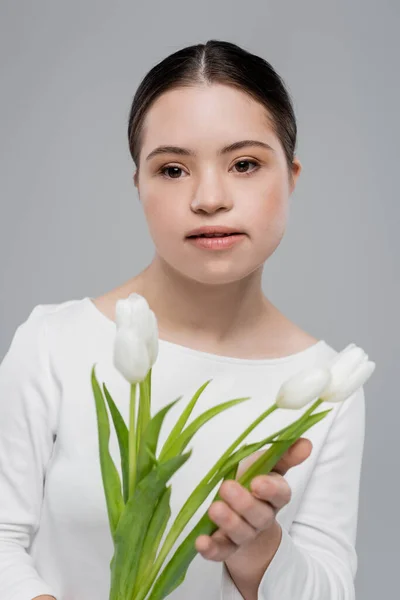 Young woman with down syndrome holding blurred tulips isolated on grey — Stock Photo