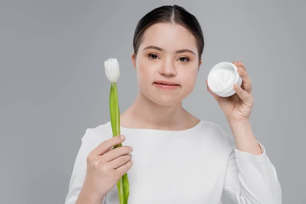 Brünette Frau mit Down-Syndrom hält Tulpe und kosmetische Creme isoliert auf grau — Stockfoto