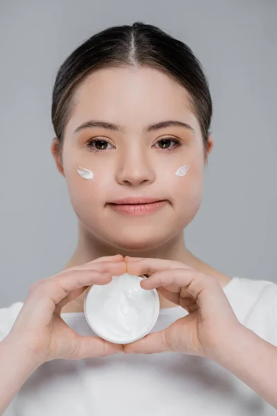 Young woman with down syndrome holding container with cosmetic cream isolated on grey — Stock Photo