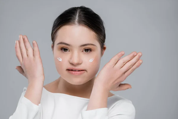 Giovane donna con sindrome di Down e crema sul viso guardando la fotocamera isolata su grigio — Foto stock