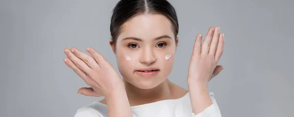 Brunette woman with down syndrome and cream on face looking at camera isolated on grey, banner — Stock Photo