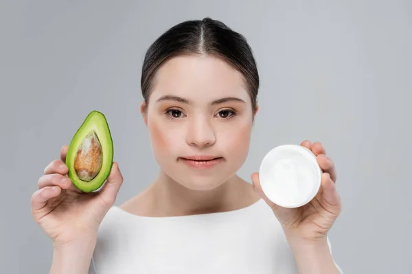 Mujer joven con síndrome de Down sosteniendo aguacate y crema cosmética aislada en gris - foto de stock