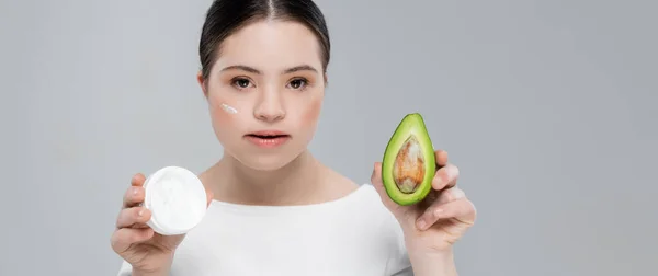 Жінка з синдромом вниз і кремом на обличчі тримає авокадо ізольовано на сірому, банер — стокове фото