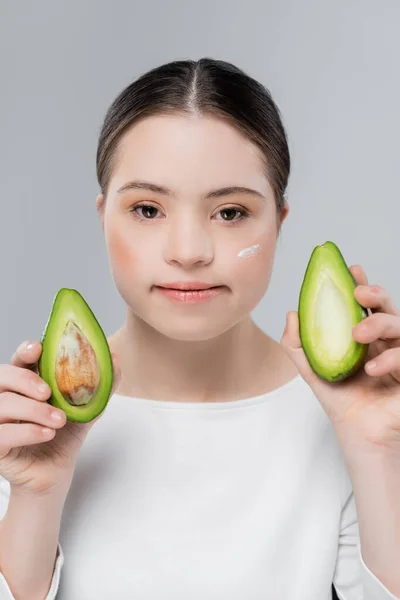 Mujer morena con síndrome de Down y crema en la cara sosteniendo aguacate orgánico aislado en gris — Stock Photo