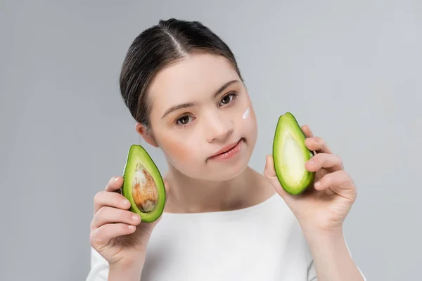 Женщина с синдромом Дауна и кремом на лице, держащая спелый авокадо изолированный на сером — стоковое фото