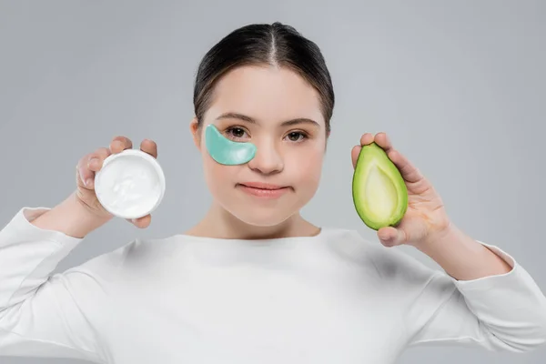 Frau mit Down-Syndrom und Augenklappe hält Avocado und Creme isoliert auf grau — Stockfoto