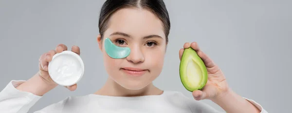 Woman with down syndrome and eye patch holding cream and avocado isolated on grey, banner — Stock Photo