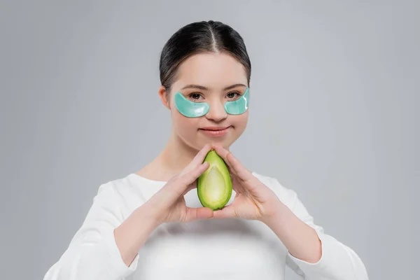 Улыбающаяся женщина с синдромом Дауна и повязками на глазу, держащая авокадо изолированным на сером — стоковое фото